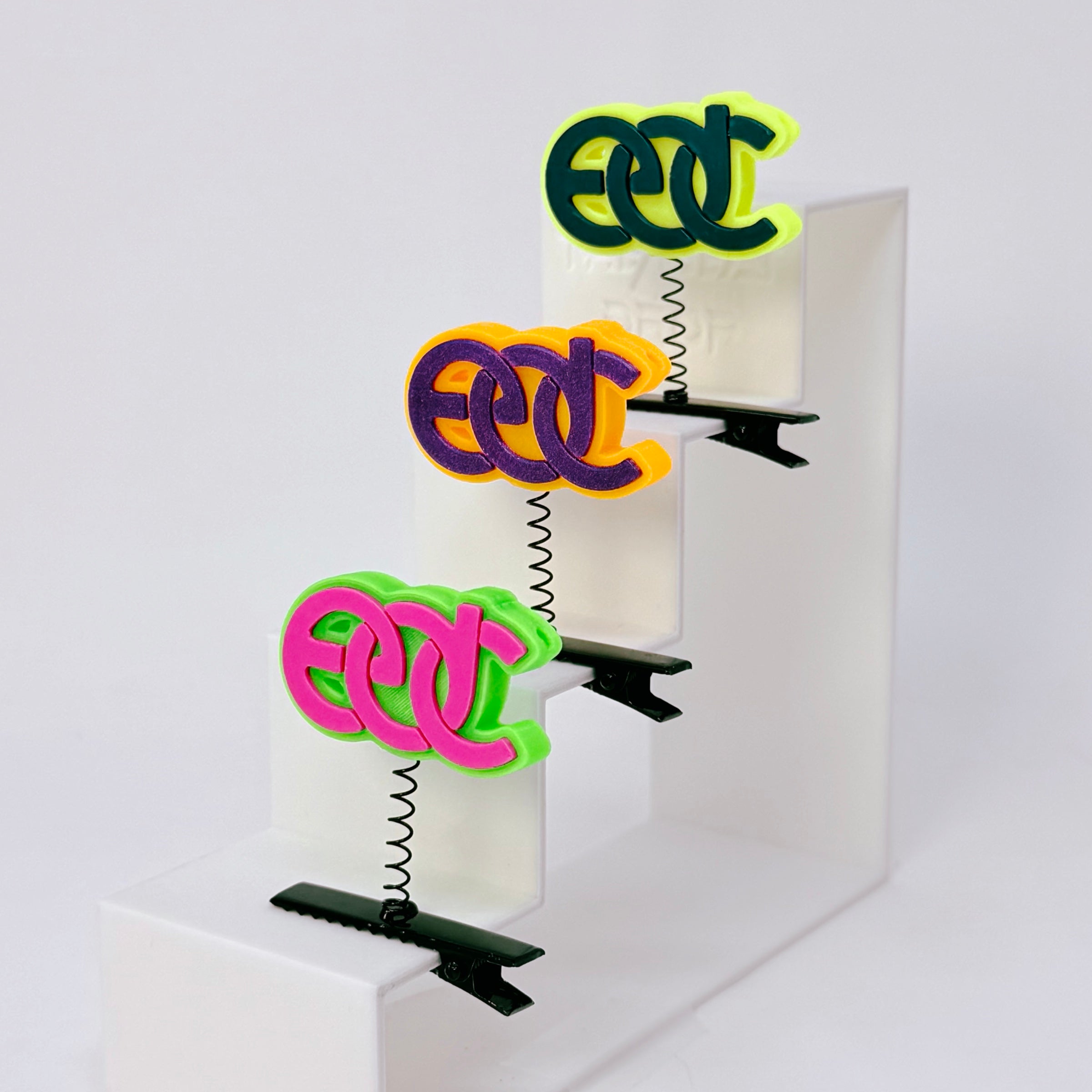 Three EDC rave sprout spring clips on white display steps — neon green hot pink, orange purple, and neon yellow turquoise colors