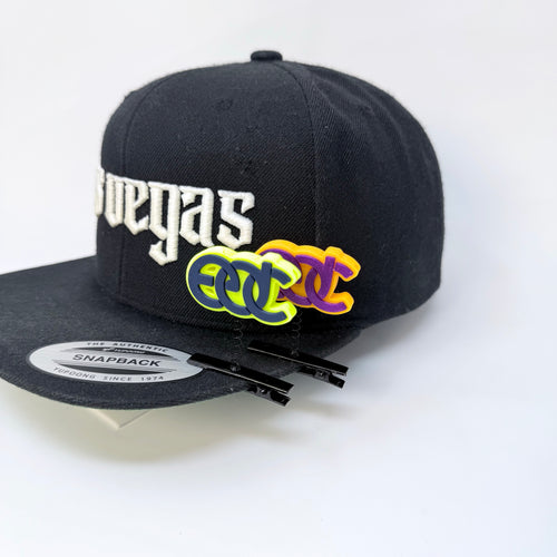 Two EDC rave sprout spring clips in orange purple and neon yellow turquoise colors on a black flat-bill hat with white Las Vegas text