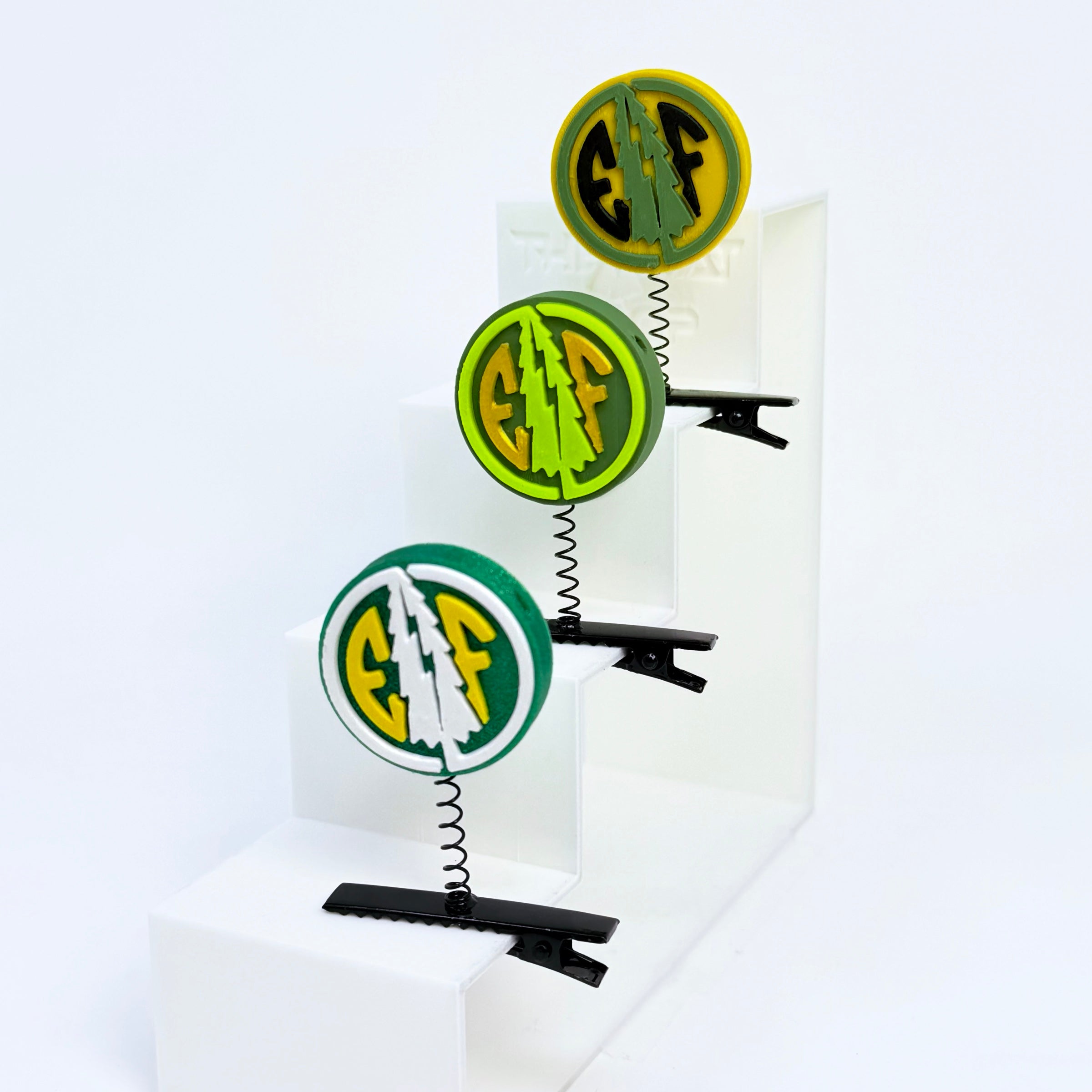 Three Electric Forest EF coin rave sprouts displayed on a white step riser in yellow/black, neon green/gold, and forest green/white colorways