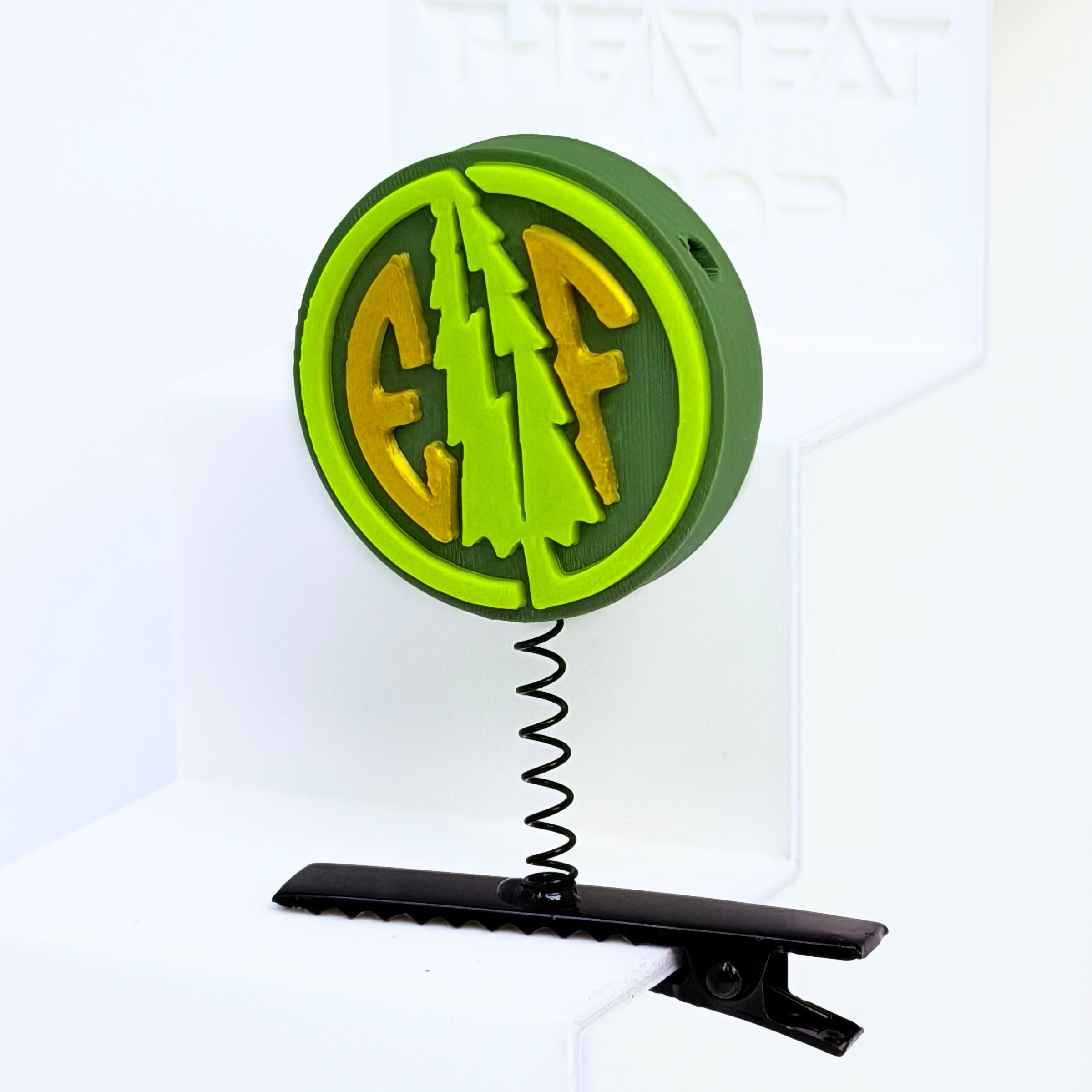 Close-up of Electric Forest EF coin rave sprout in dark olive green with neon green pine tree and gold EF letters, mounted on black metal spring hair clip