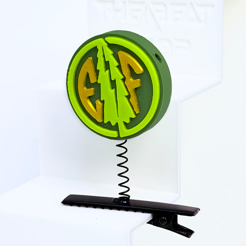 Close-up of Electric Forest EF coin rave sprout in dark olive green with neon green pine tree and gold EF letters, mounted on black metal spring hair clip
