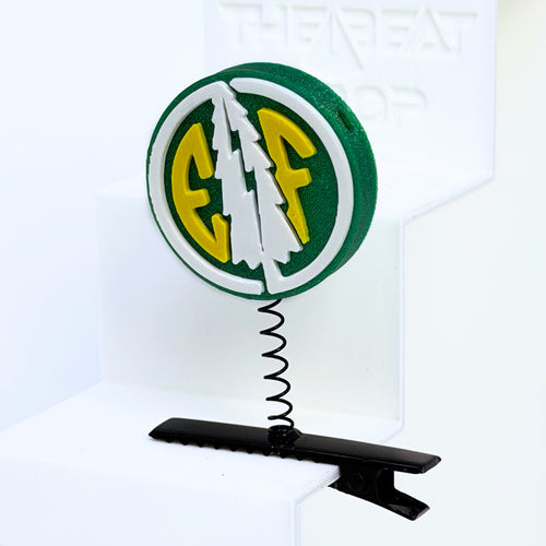 Close-up of Electric Forest EF coin rave sprout in forest green with white pine tree and yellow EF letters, mounted on black metal spring hair clip