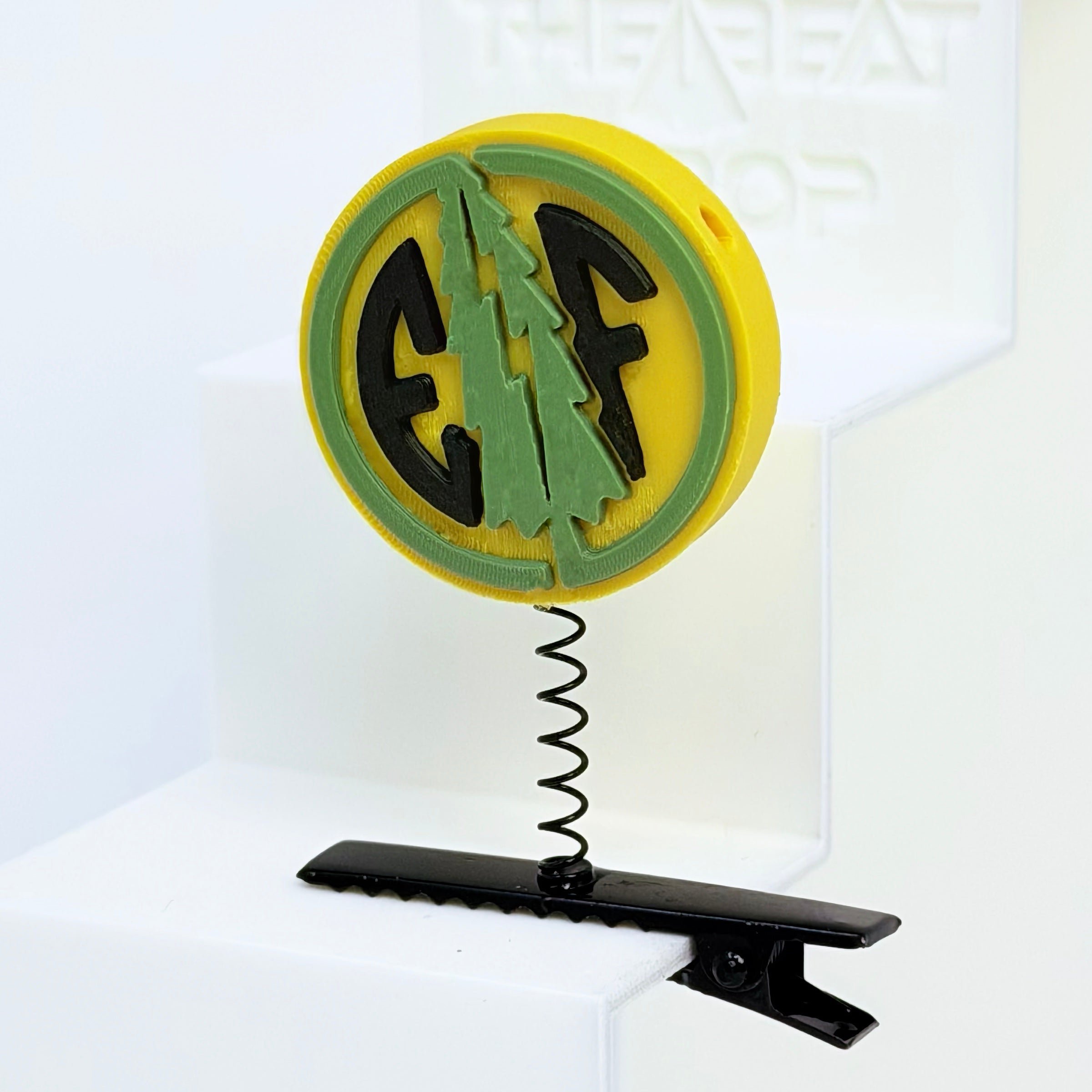 Close-up of Electric Forest EF coin rave sprout in bright yellow with sage green pine tree and black EF letters, mounted on black metal spring hair clip