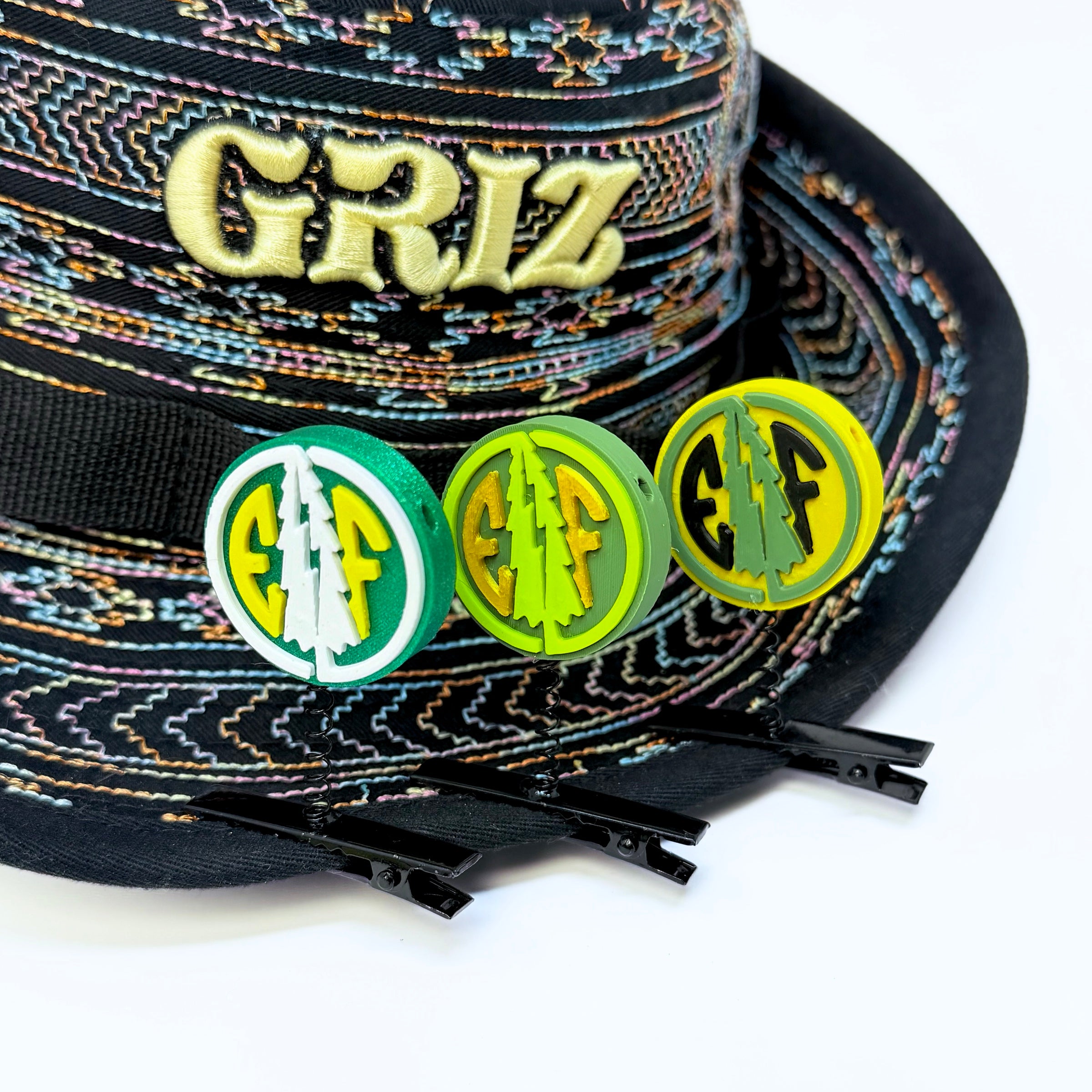 Three Electric Forest EF coin rave sprouts in green/white/yellow, olive/neon green/gold, and yellow/sage/black colorways clipped to the brim of a GRiZ festival bucket hat