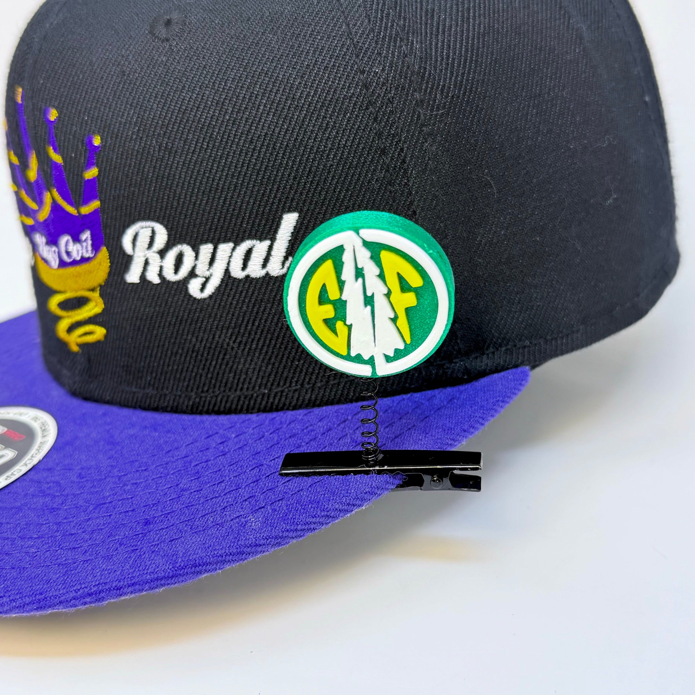 Electric Forest EF coin rave sprout in forest green, white, and yellow clipped to the panel of a black and purple Royal snapback hat