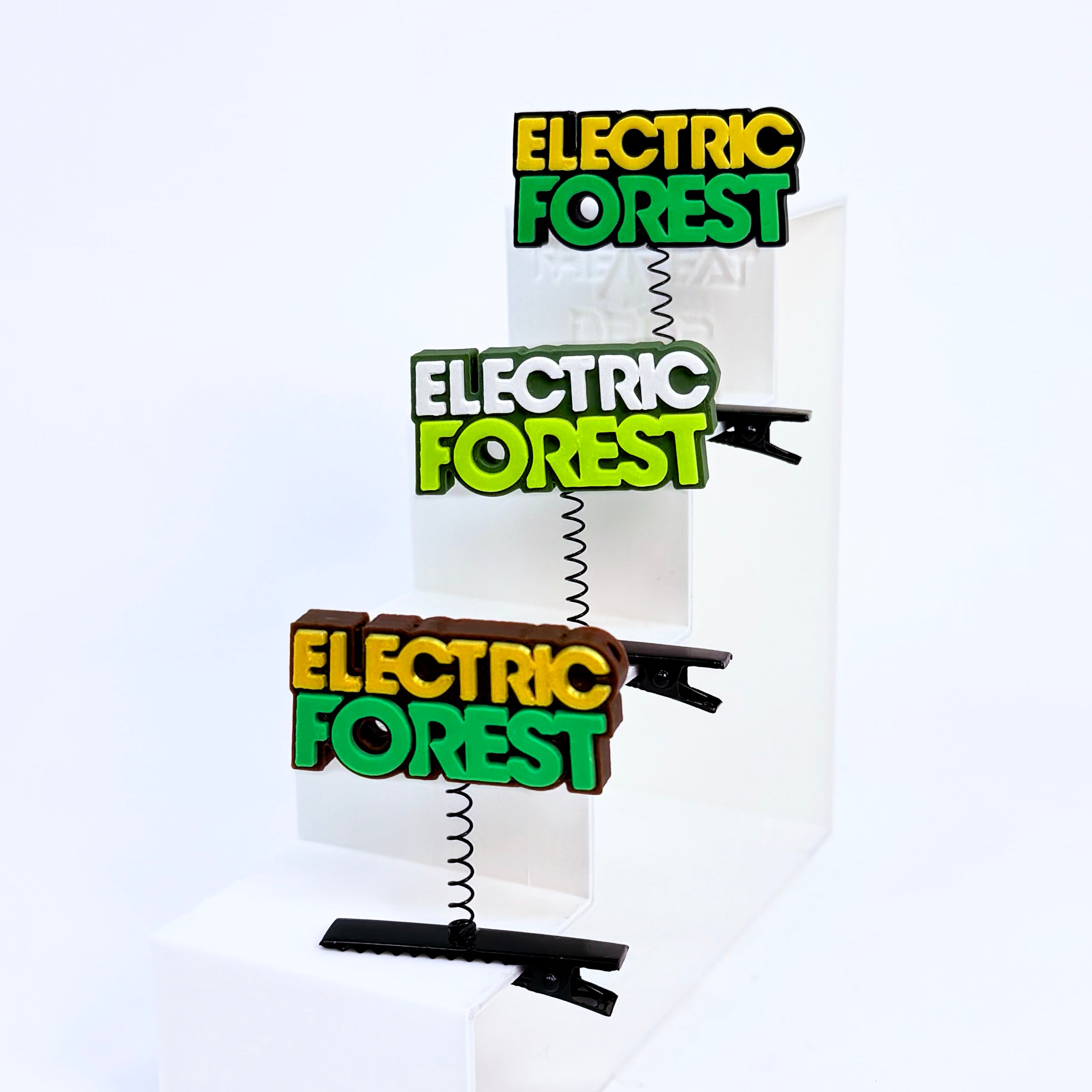 Three Electric Forest text rave sprouts displayed on a white step riser in yellow/black, dark green/white/neon green, and brown/gold/green colorways