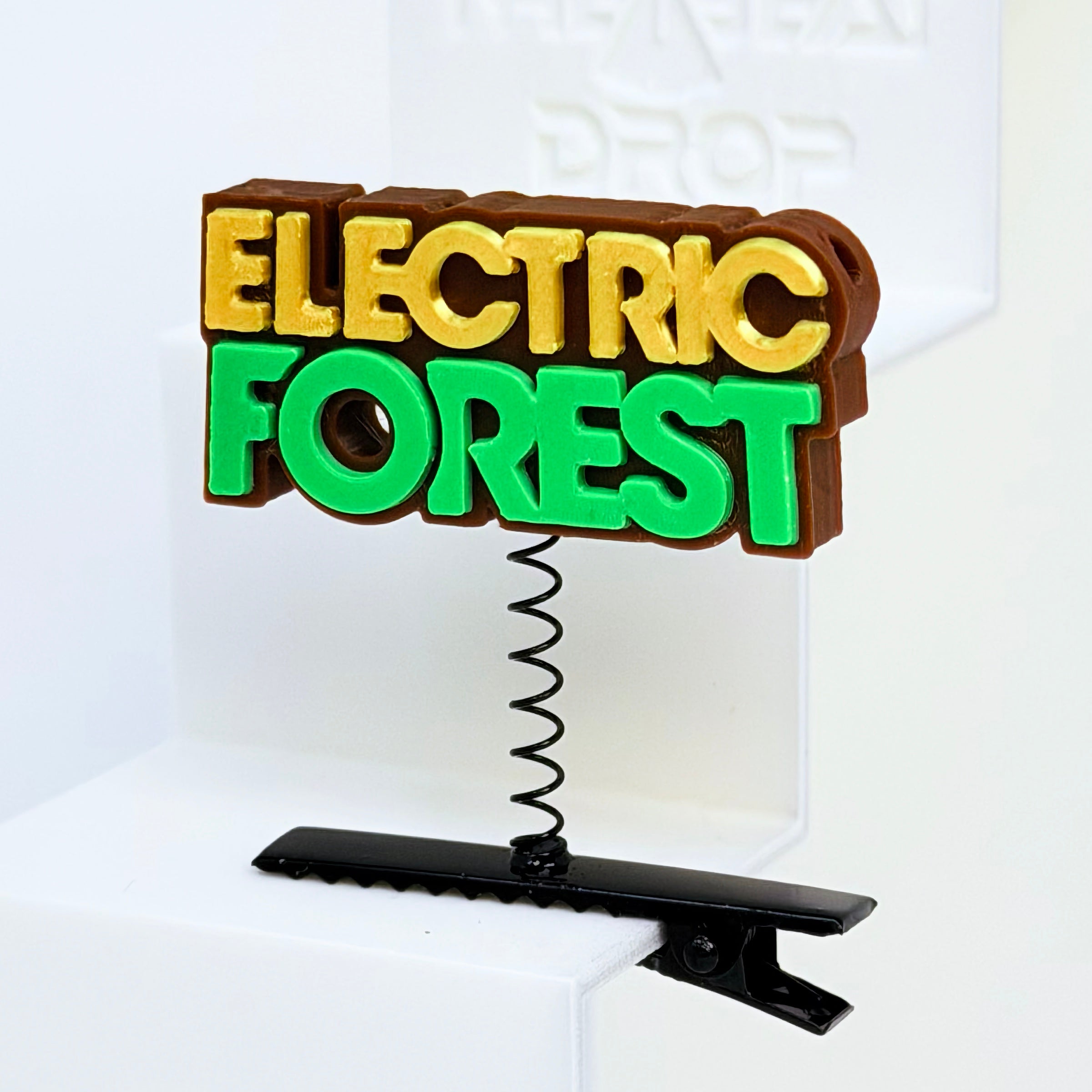 Close-up of Electric Forest text rave sprout with brown base, gold "ELECTRIC" lettering, and bright green "FOREST" lettering, mounted on black metal spring hair clip