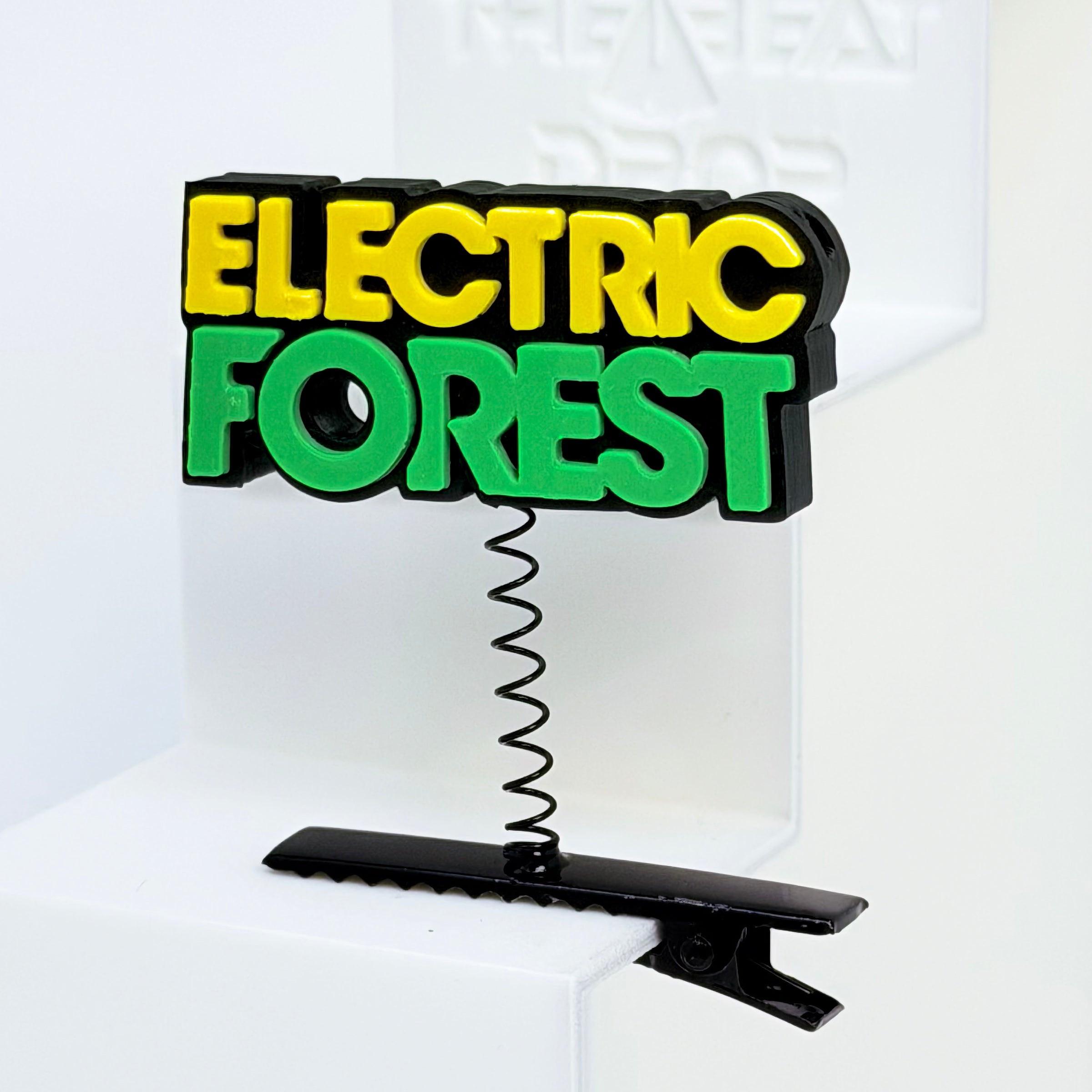 Close-up of Electric Forest text rave sprout with black base, yellow "ELECTRIC" lettering, and bright green "FOREST" lettering, mounted on black metal spring hair clip