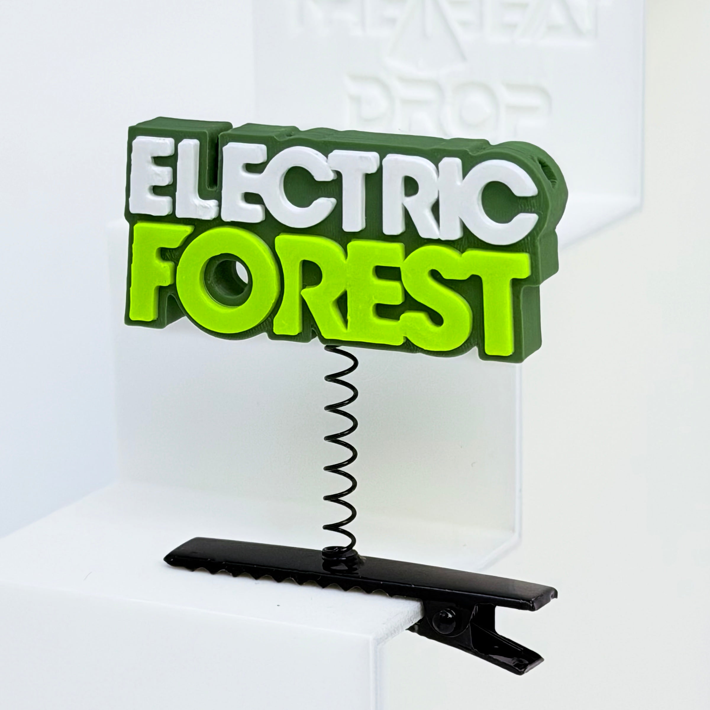 Close-up of Electric Forest text rave sprout with dark olive green base, white "ELECTRIC" lettering, and neon yellow-green "FOREST" lettering, mounted on black metal spring hair clip