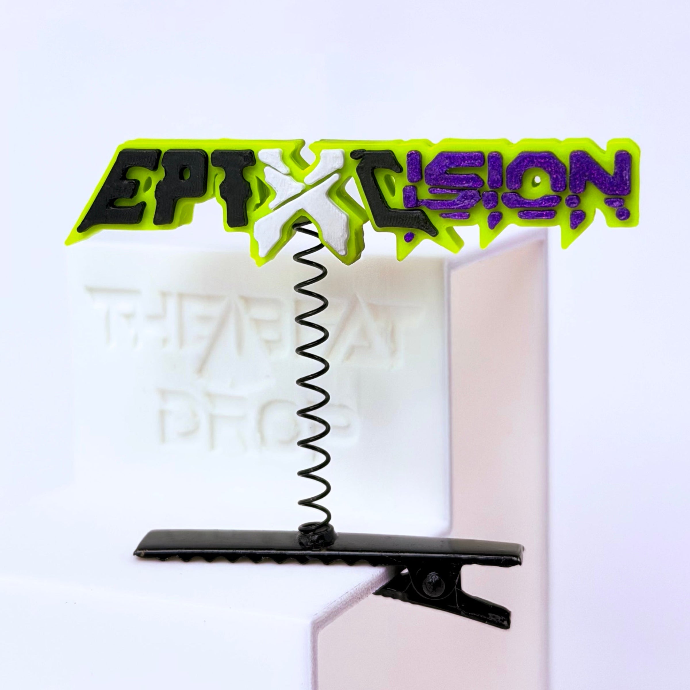 A black hair clip with a coiled spring and a colorful Excision and Eptic graphic topper, displayed on a white block against a light background.