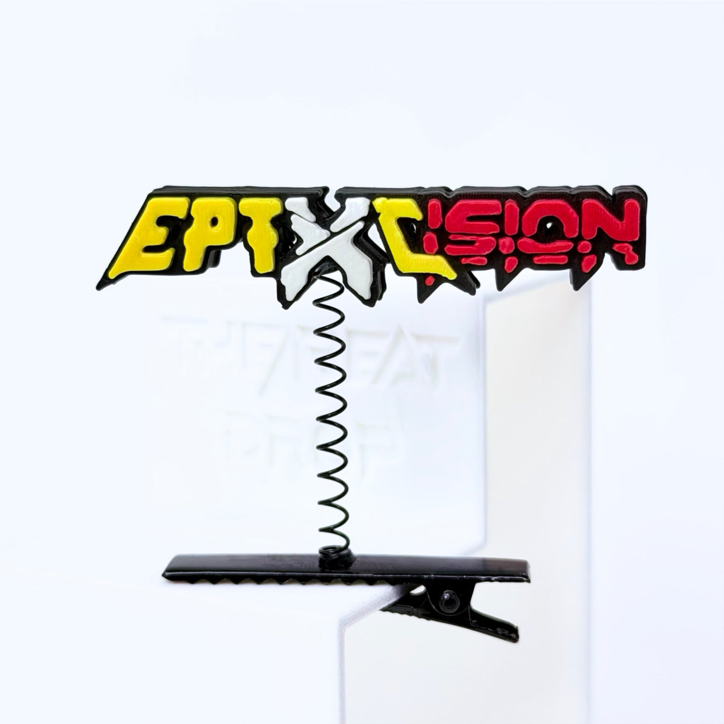 A novelty hair clip featuring the stylized text Excision and Eptic in yellow, white, and red with a black outline, suspended on a black spring from a black clip, shown on a white background.