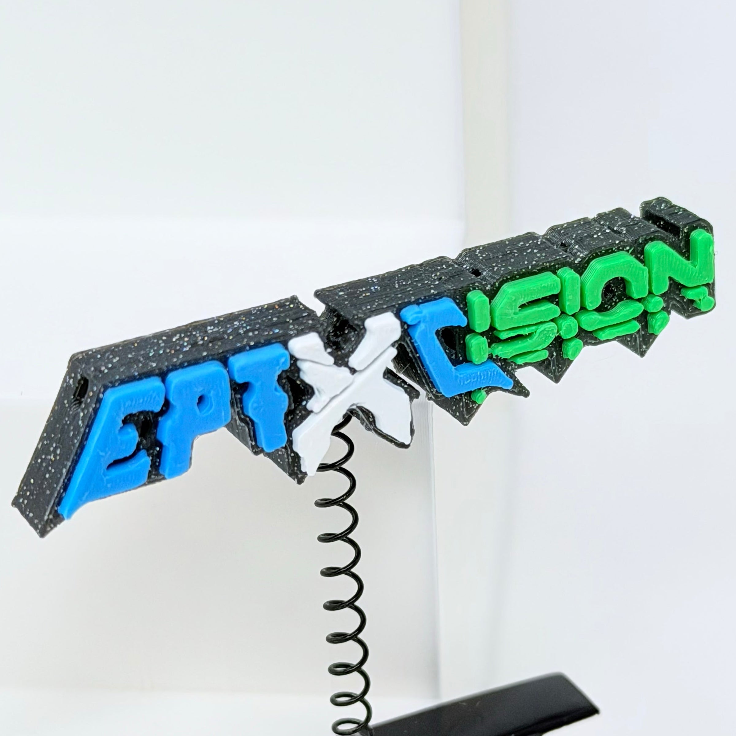 A 3D-printed clip-on accessory featuring the text Excision and Eptic in stylized blue, white, and green lettering on a black glittery background, mounted on a black spring clip.