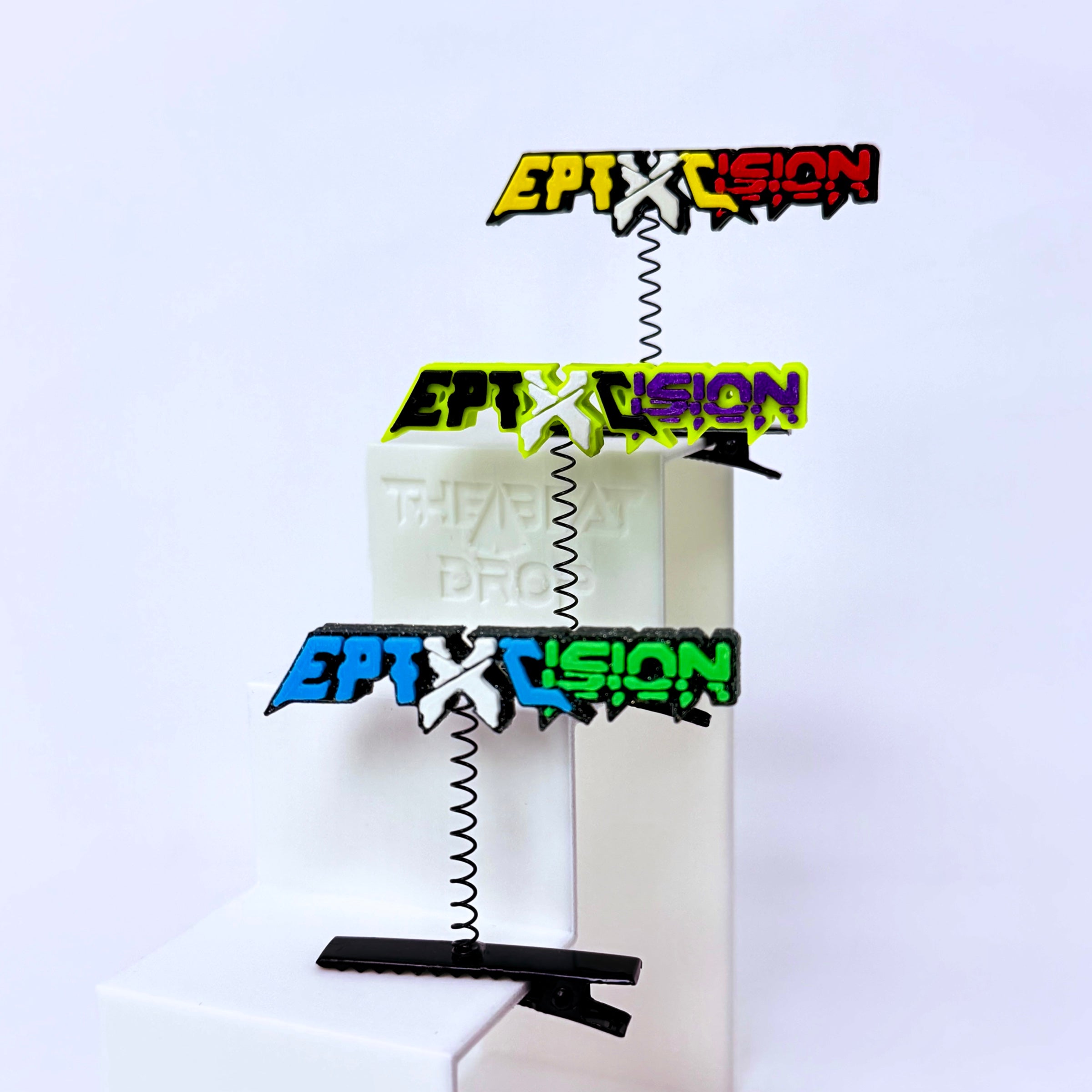 Three colorful Excision and Eptic logo spring clips in yellow, green, and blue, stacked vertically on a white display stand.