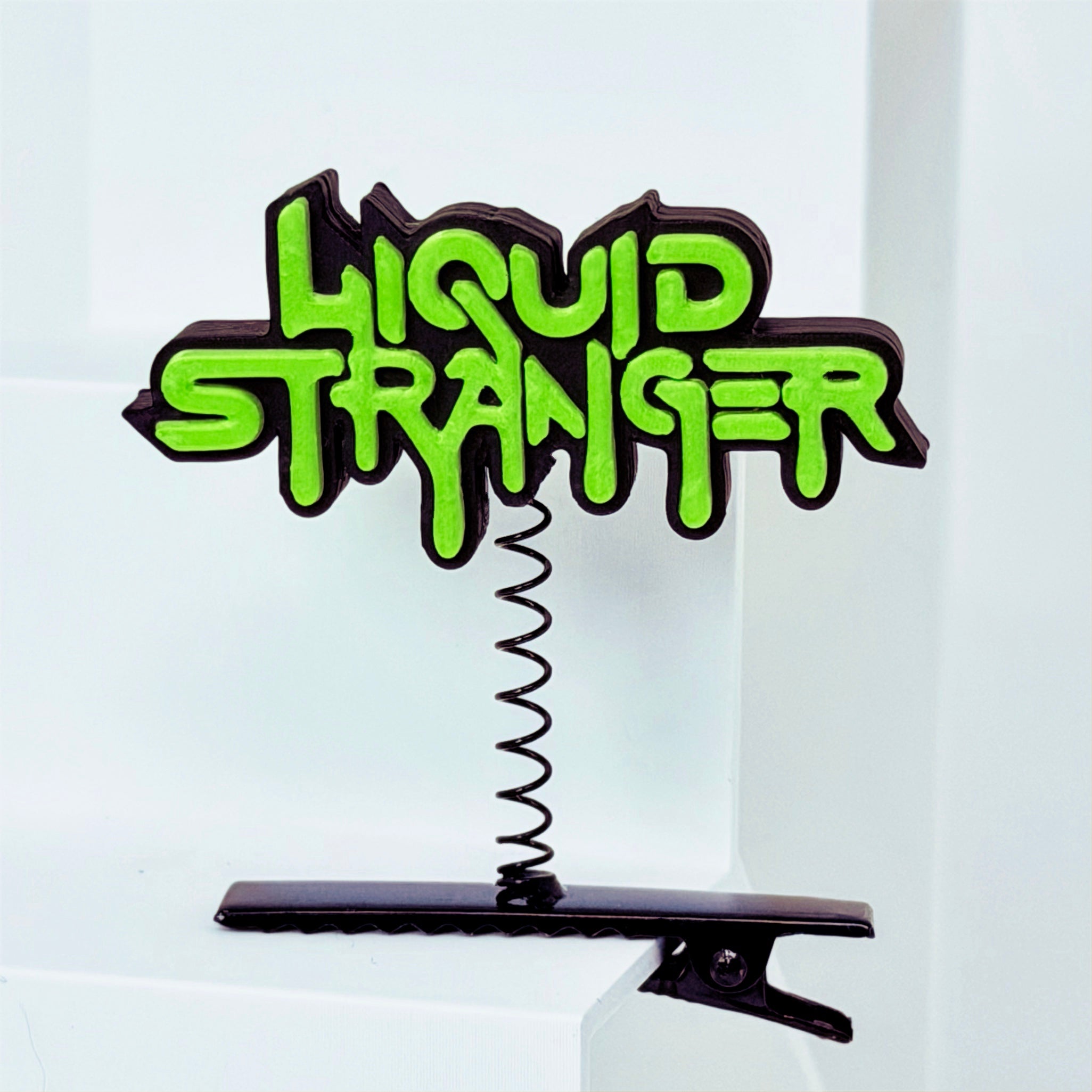 A black rave sprout hair clip featuring LIQUID STRANGER text in bright neon green graffiti-style lettering, mounted on a black spring-loaded clip.