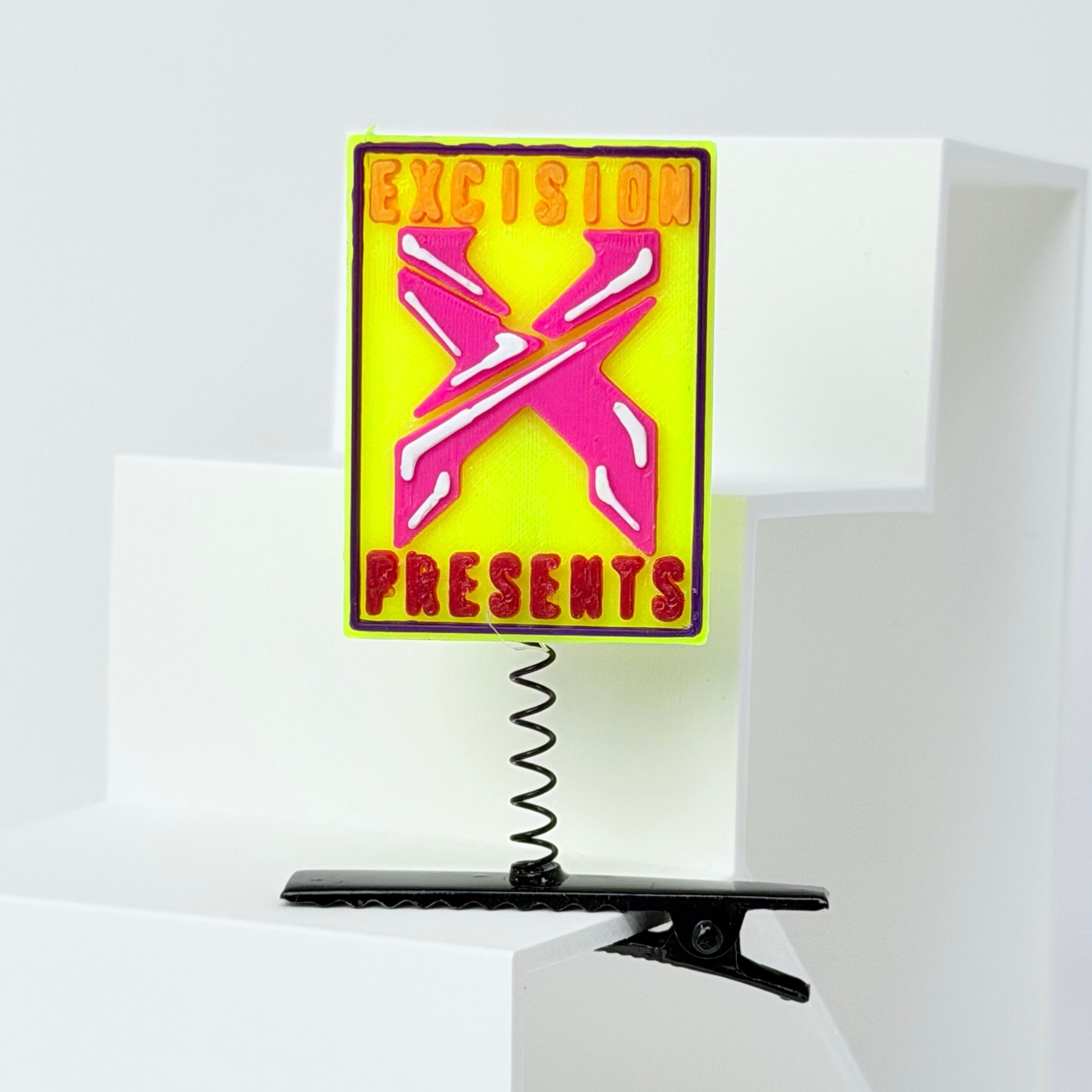 A novelty sign featuring a bright yellow background, a pink 3D-effect 'X' in the center, and the words 'EXCISION PRESENTS' in red and orange lettering. The sign is mounted on a black metal spring clip, displayed on a white geometric riser against a light gray background.