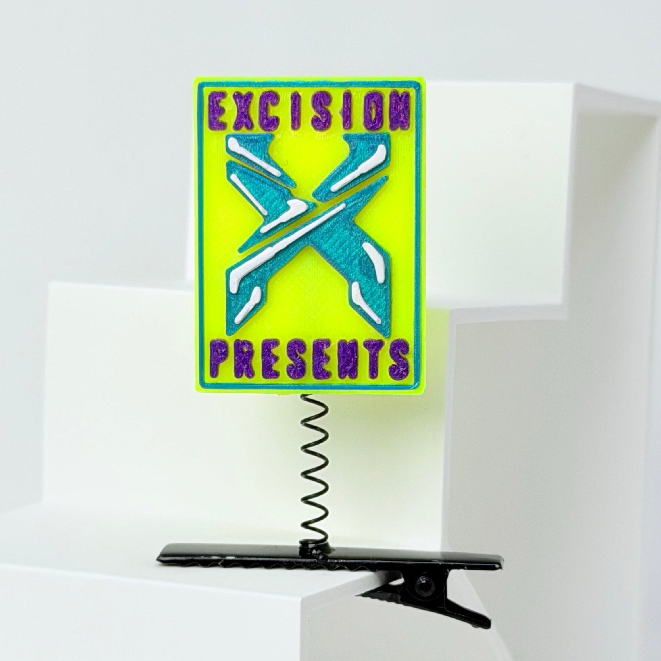 A rectangular neon yellow sign with a green border, featuring a stylized blue and white 'X' and the words 'EXCISION PRESENTS' in purple. The sign is mounted on a black spring clip attached to a white geometric display stand.