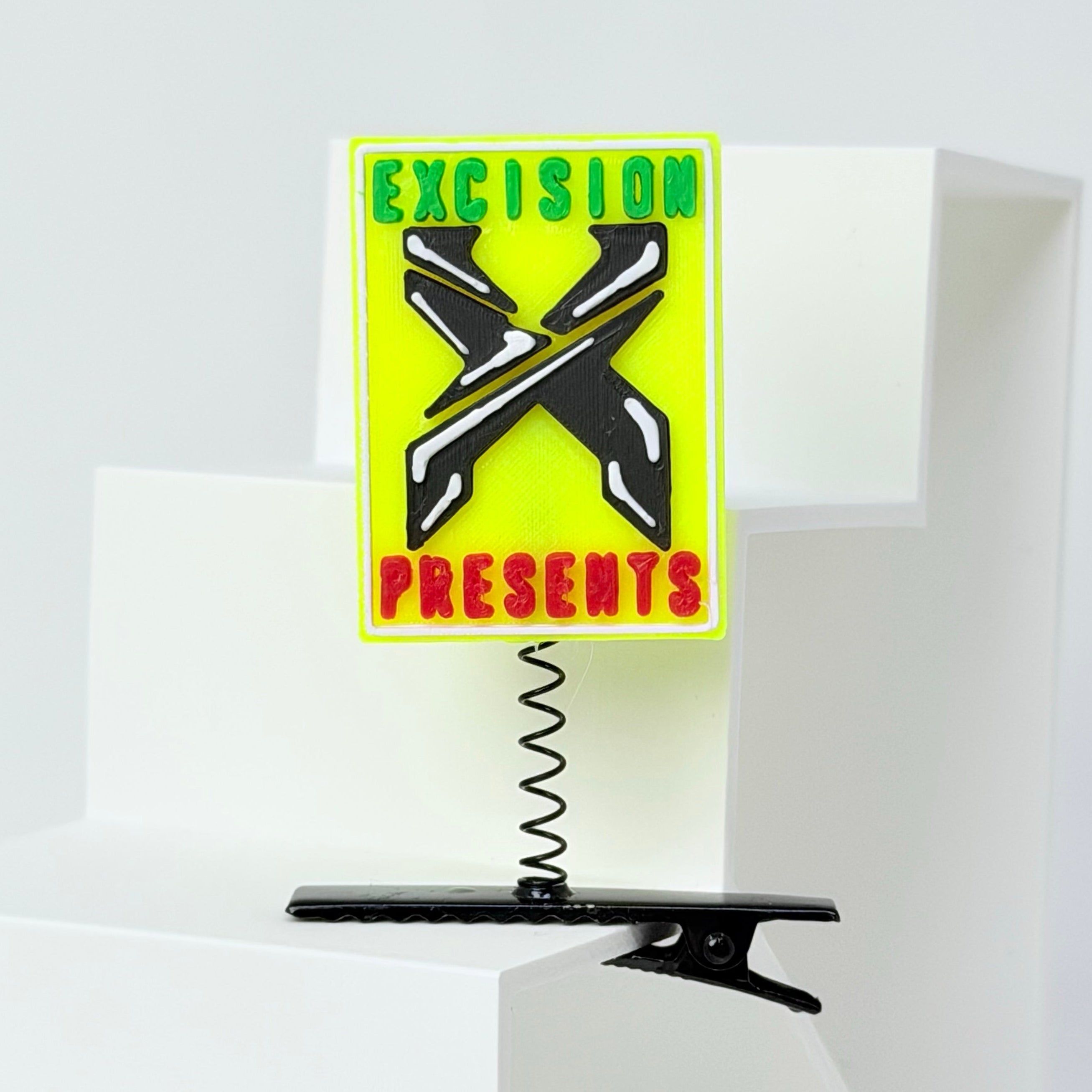 A rectangular yellow sign with a black crossed-X logo and the words 'EXCISION PRESENTS' in green and red, mounted on a black spring clip stand against a white geometric display.