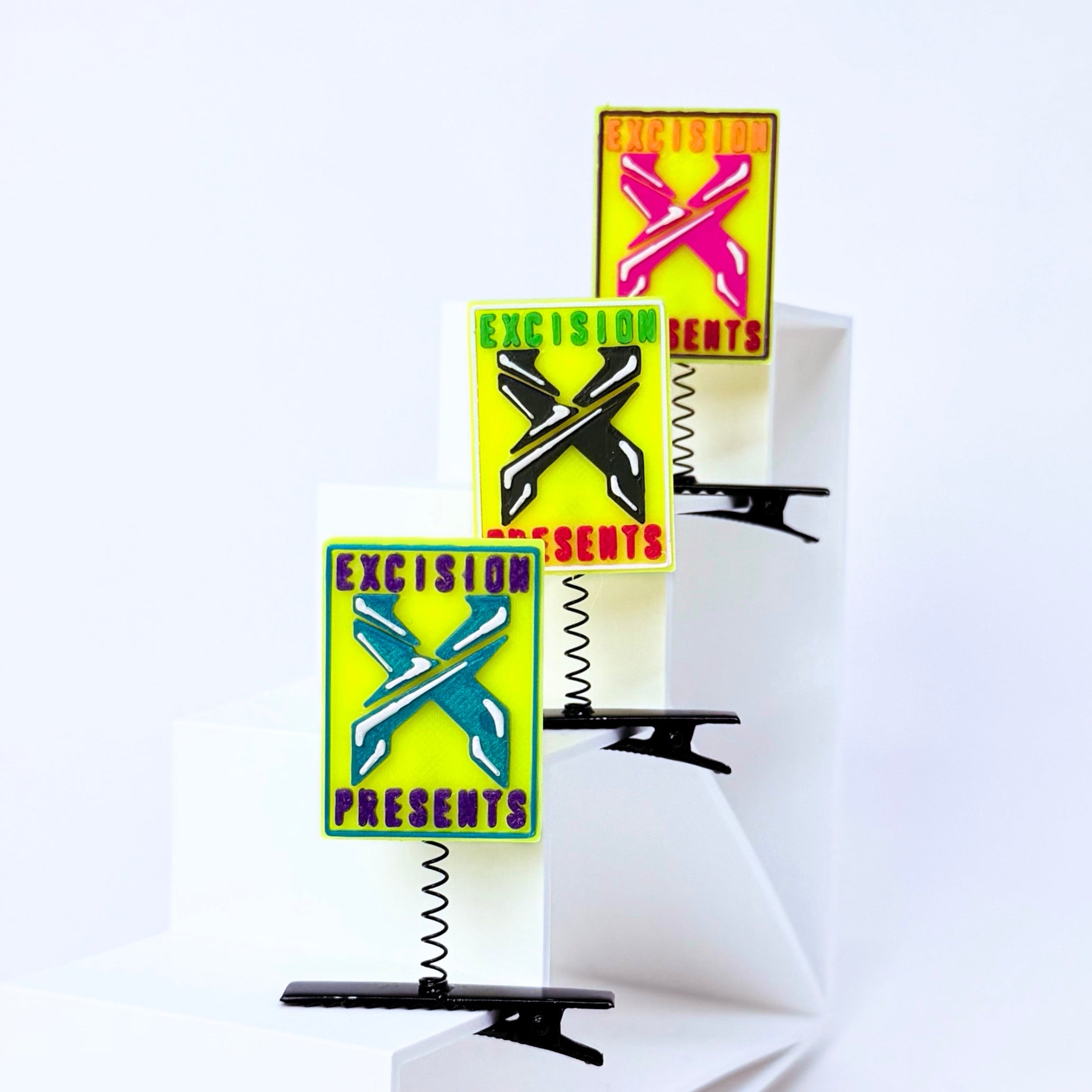 Three rectangular enamel pins with spring clips are displayed on white geometric risers. Each pin features a bright yellow-green background with a large crossed-color 'X' graphic and the text 'EXCISION PRESENTS' in purple.
