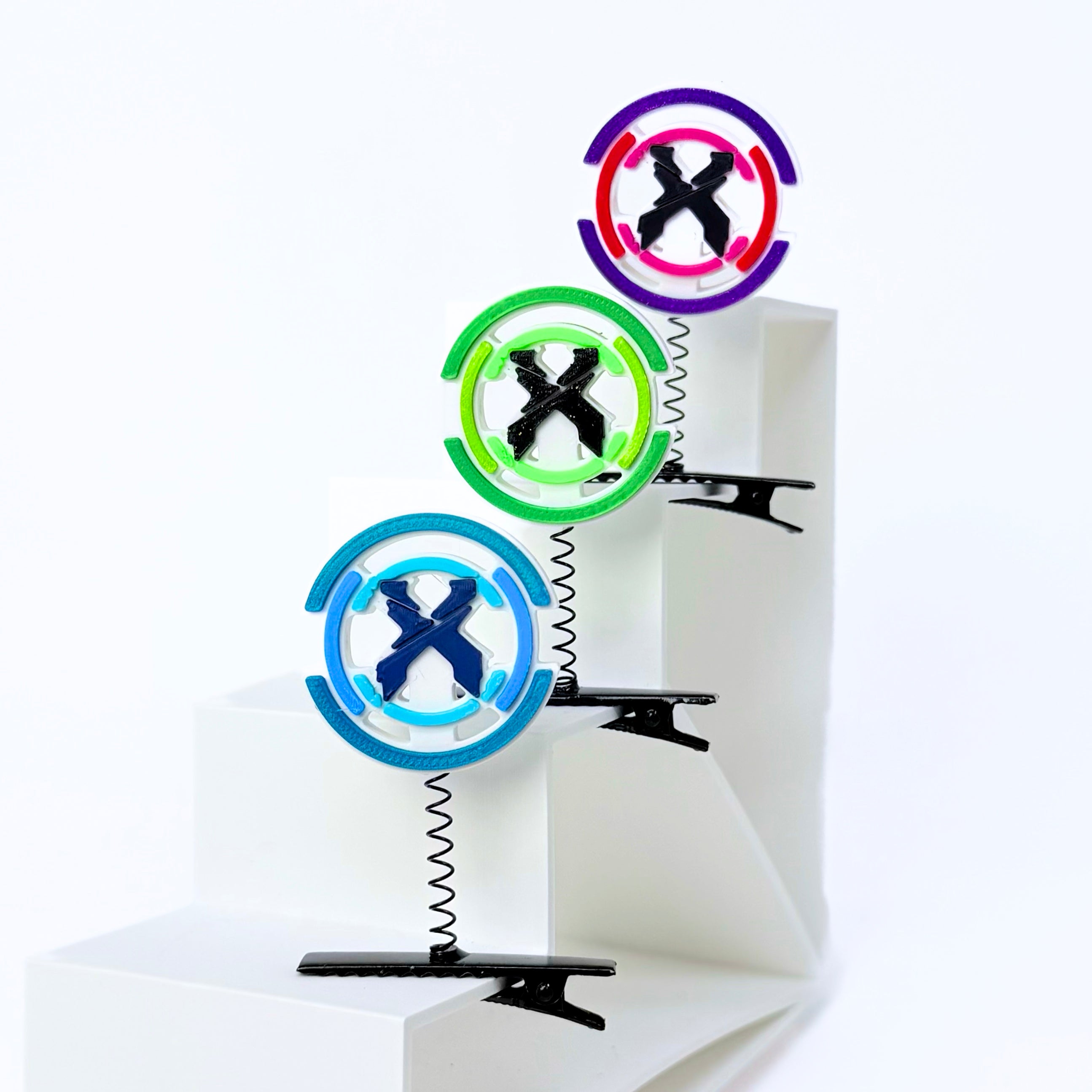 Three colorful circular spring-mounted clip stands arranged on white geometric steps against a white background. The stands feature black spring coils and black plastic clips at the bottom, with circular tops in purple, green, and blue containing black X center designs.