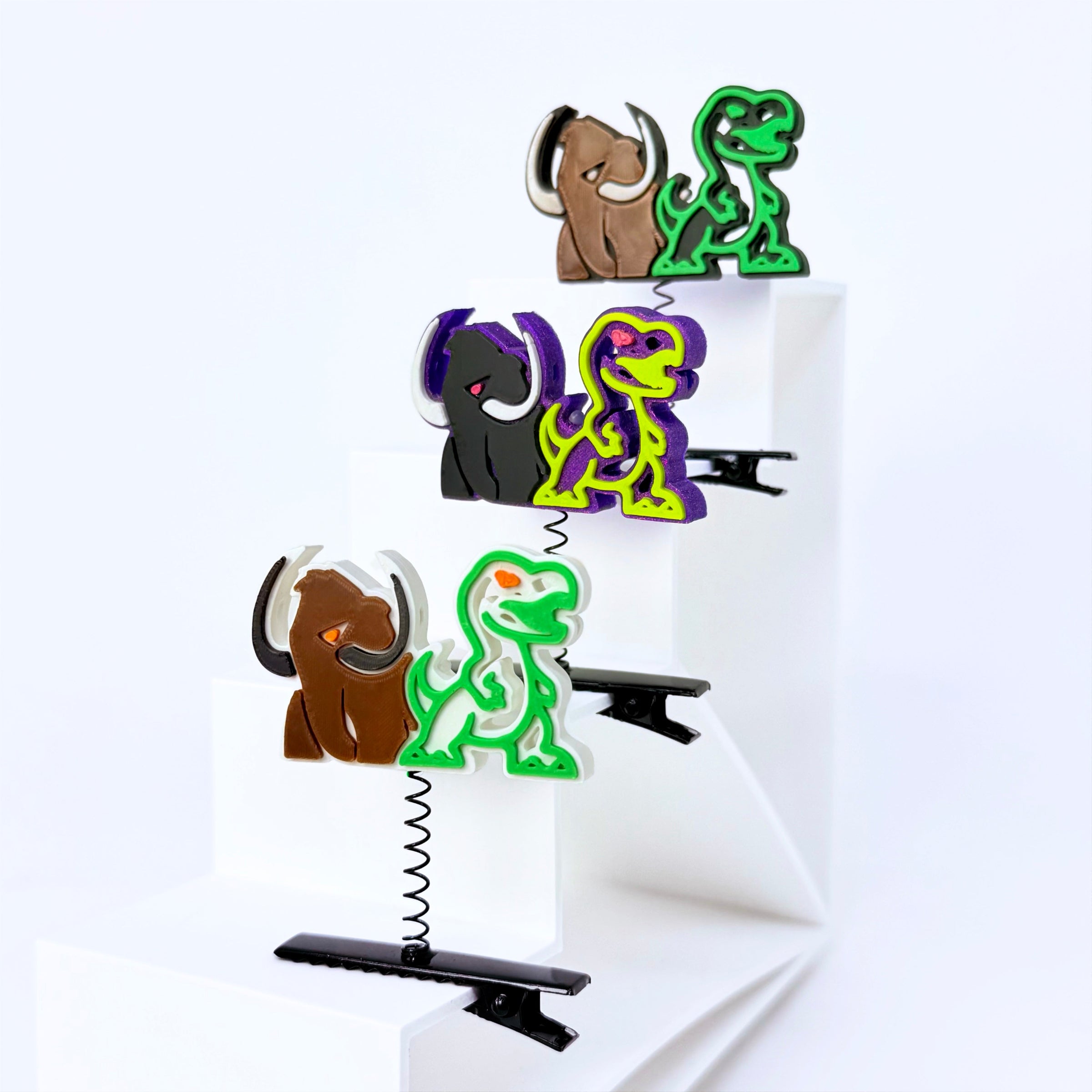 Three dinosaur-shaped hair clips in brown, black, and green, each attached to a black spring clip and displayed on a white geometric riser against a white background.