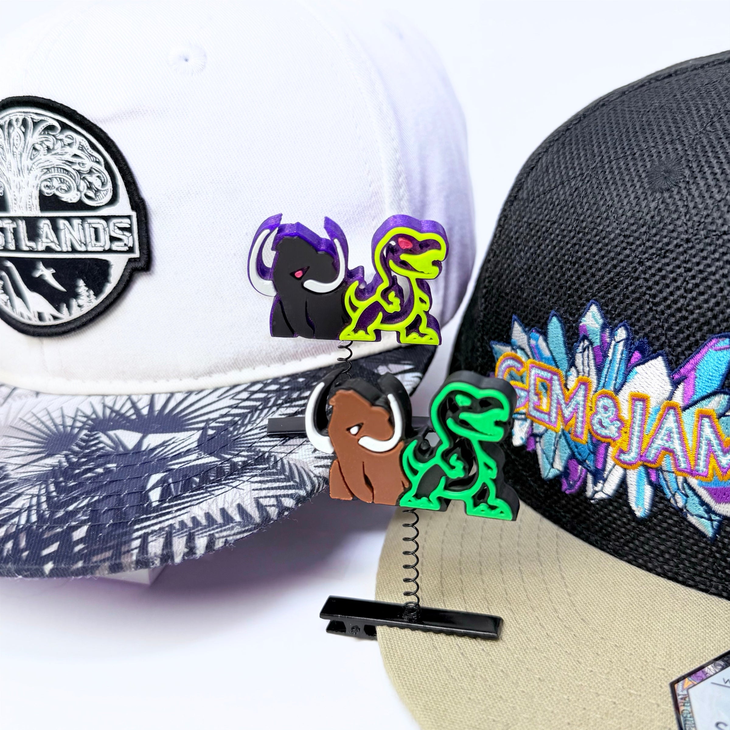 Three colorful dinosaur-shaped spring clips are displayed on three different baseball caps, including a white cap with a black patch, a black cap with a colorful 'Gem of the Sun' patch, and a beige cap with a palm leaf pattern.