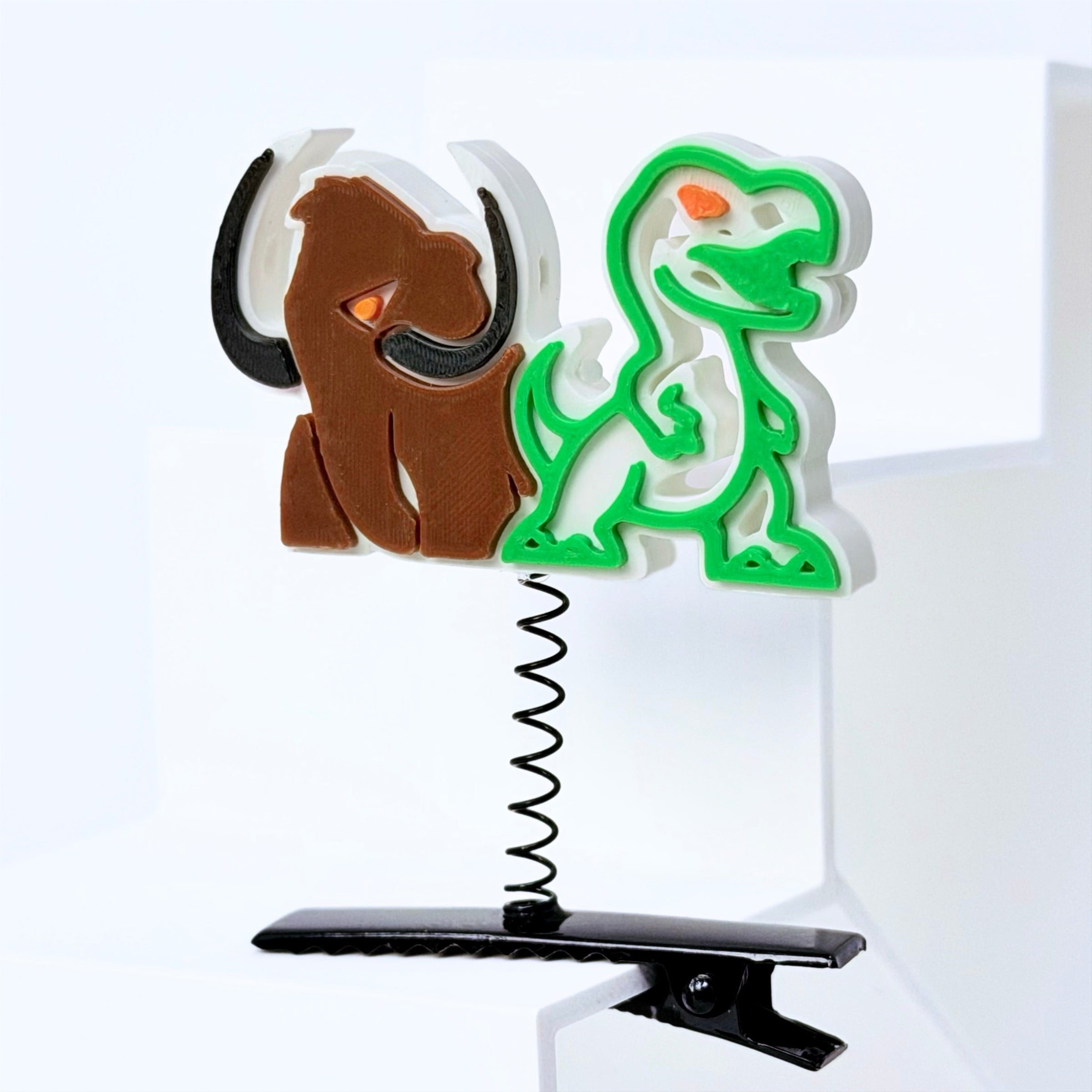A novelty hair clip featuring a spring-loaded design with a brown mammoth and a green T-Rex figure, mounted on a black spring and clip base.