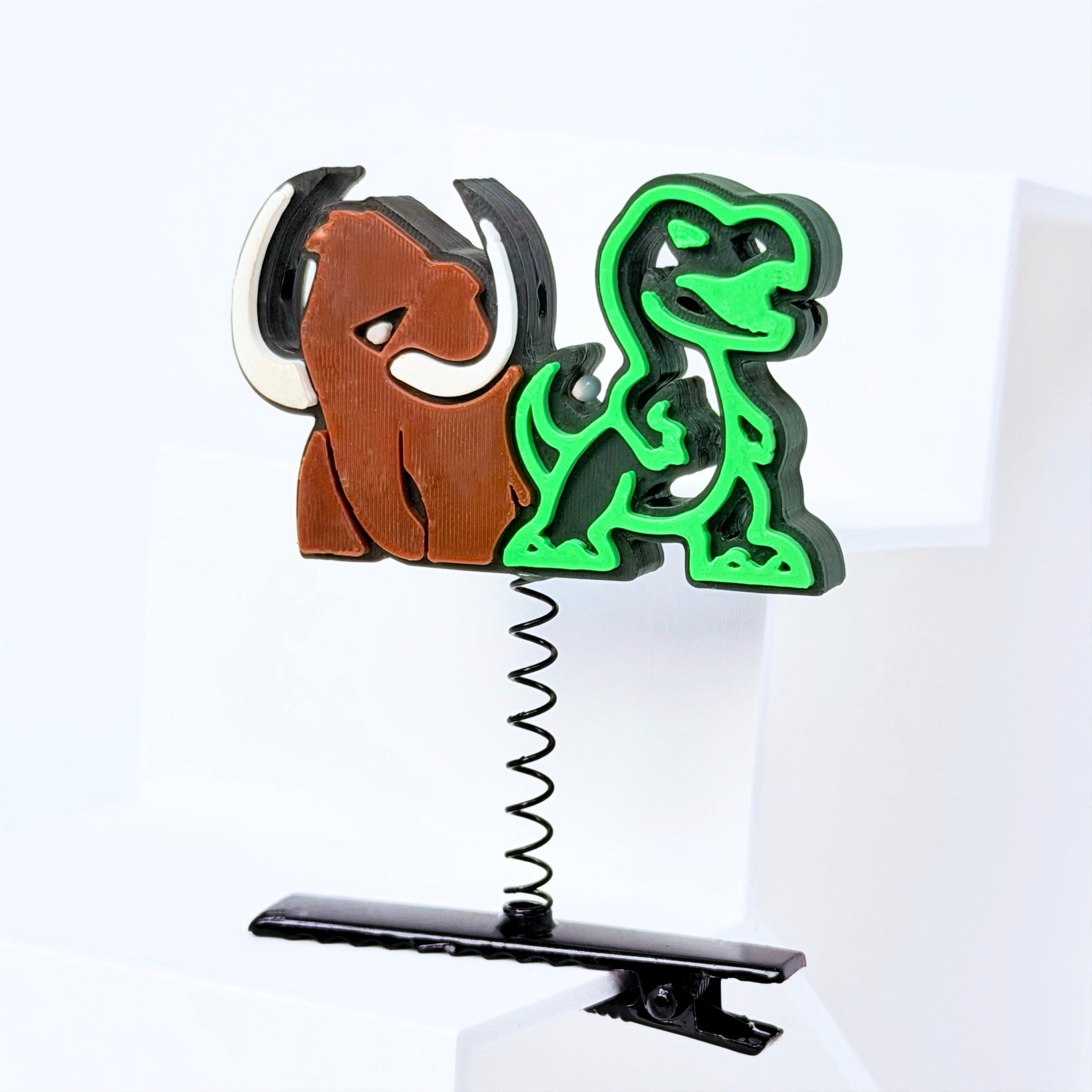 A novelty desk accessory featuring a wooden mammoth and a green plastic dinosaur cutout mounted on a black metal spring and clip base, set against a clean white background.