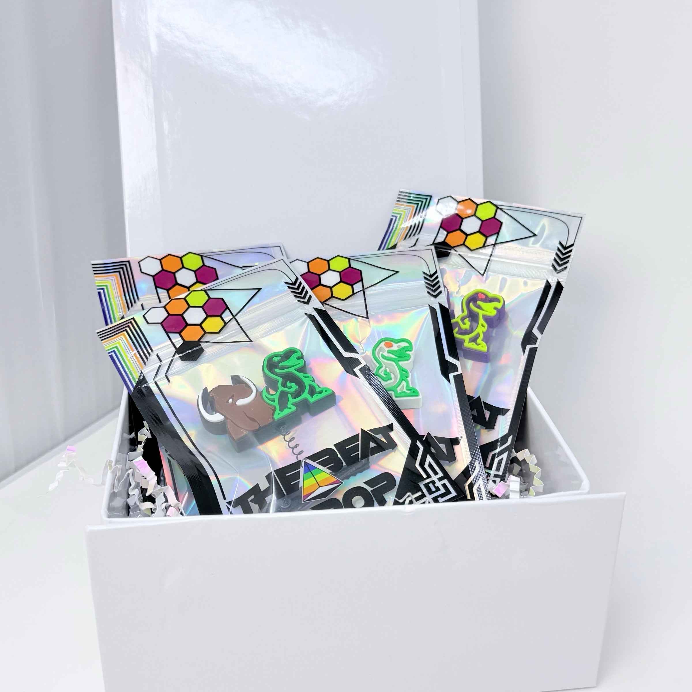 A white gift box filled with multiple holographic zip pouches decorated with geometric patterns, cartoon animal graphics, and black geometric borders, arranged on a white surface against a white background.