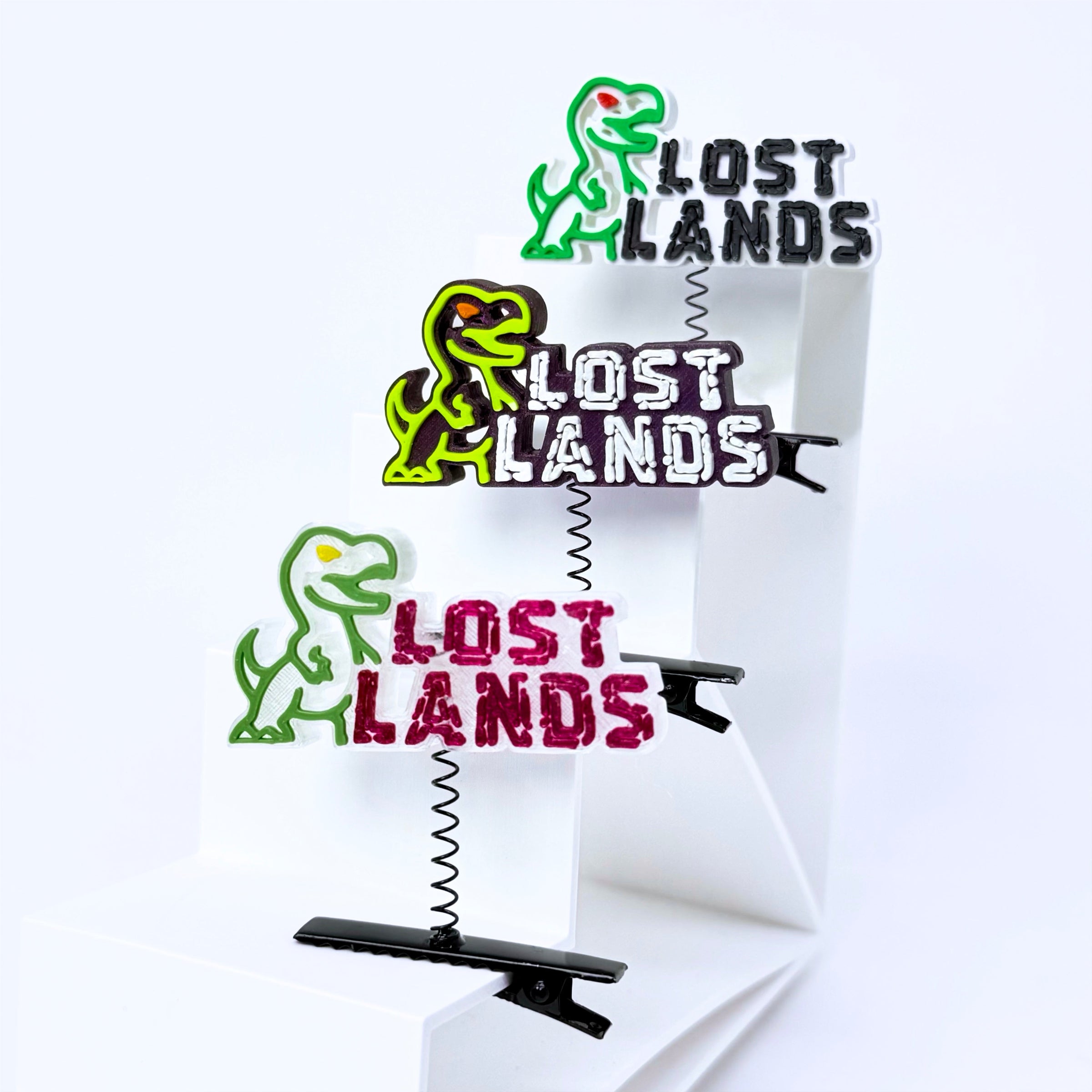 Three T-Rex clip-on hair clips featuring the 'Lost Lands' logo, displayed on a white geometric stand against a white background.