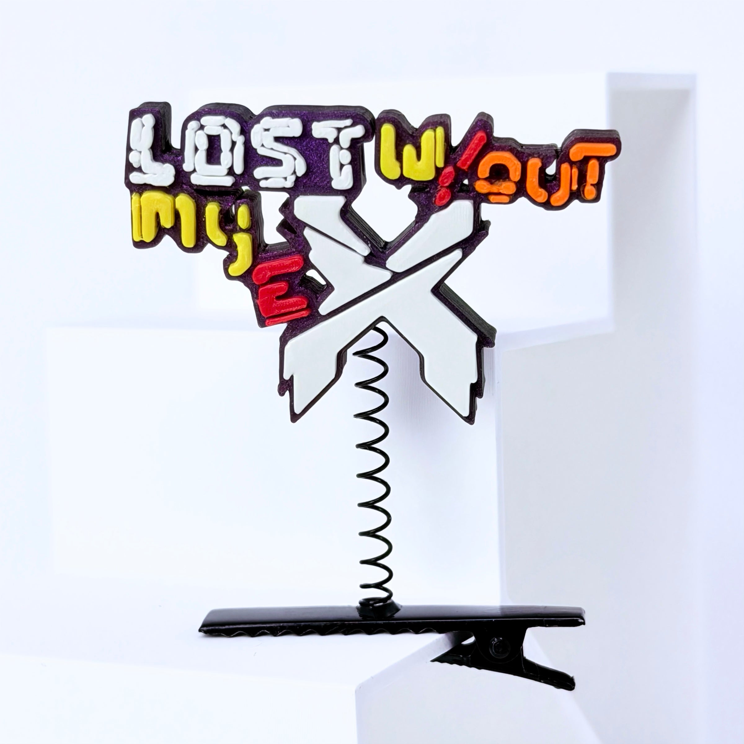 A novelty hair clip standee featuring the text 'LOST WITHOUT MY EX' in a graffiti-style multicolor font with a large white 'X' in the center, mounted on a black spring and black clip base.