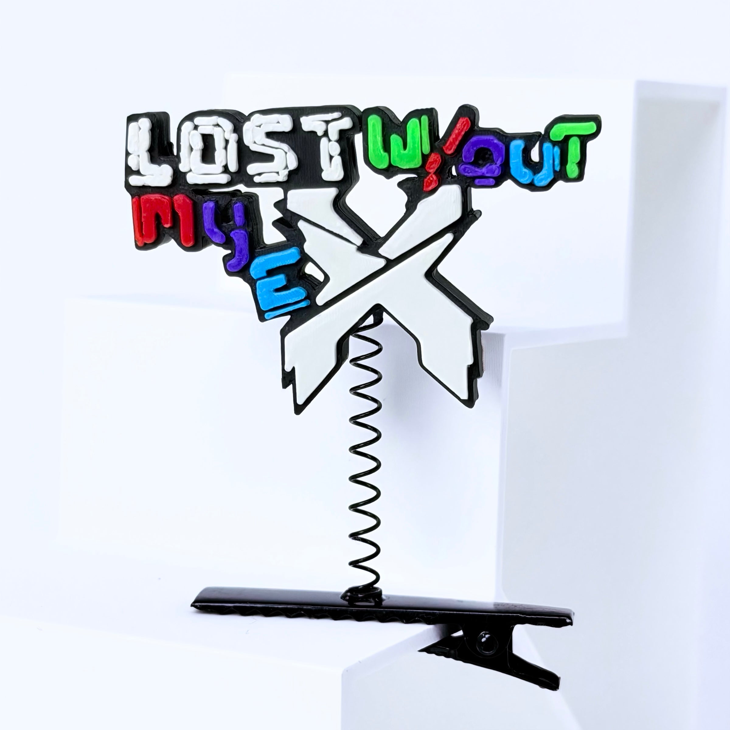 A novelty hair accessory featuring a black metal clip at the bottom and a spring-loaded top with the text 'LOST WITHOUT MY EX' in colorful graffiti-style lettering, shown on a white geometric background.