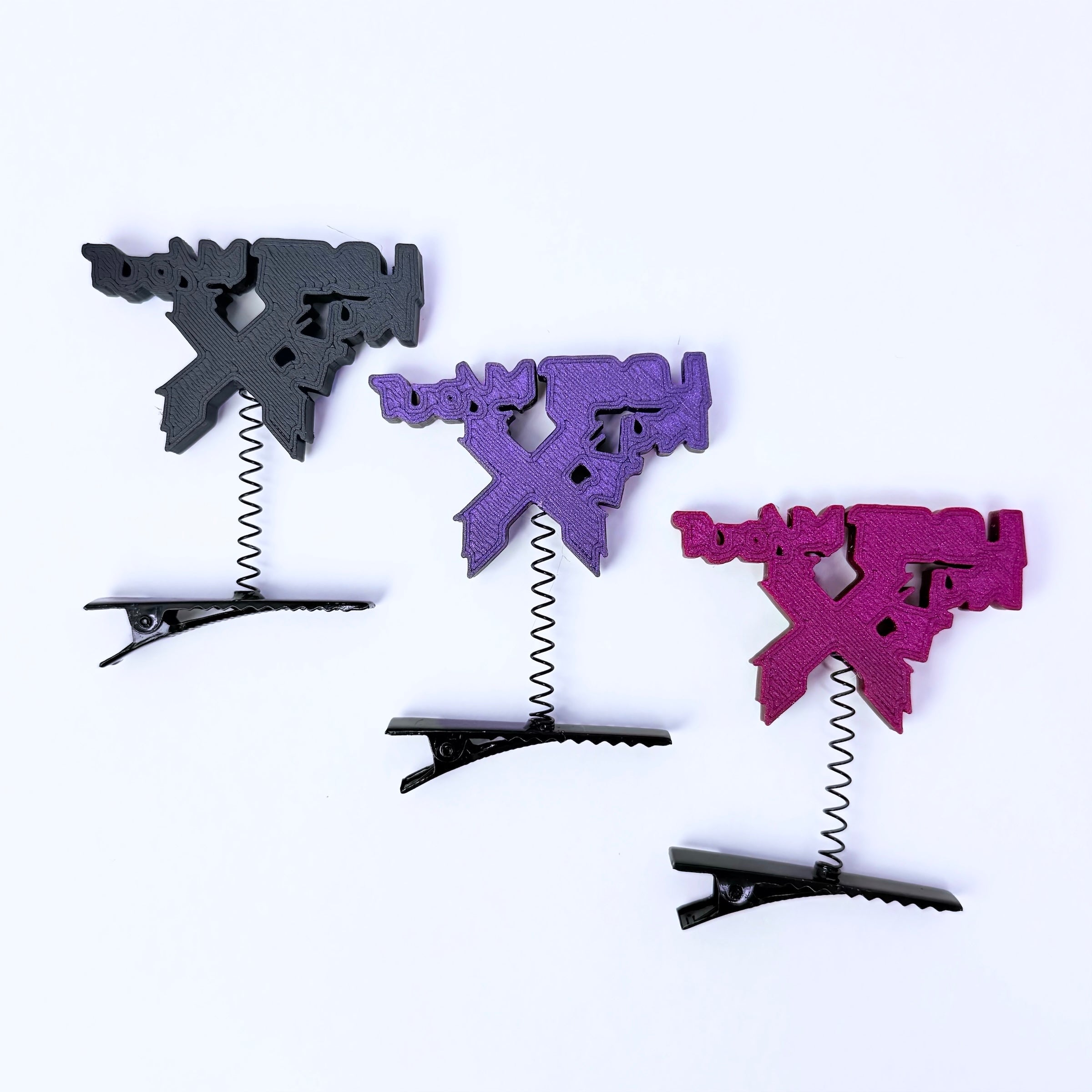 Three textured letter 'X' hair clips in black, purple, and magenta, each with a coiled spring and a black metal spring clip at the bottom, arranged diagonally on a white background.